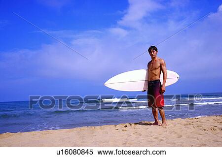 Stock Photography - Man holding the surfboard on the beach and looking at the camera. Fotosearch