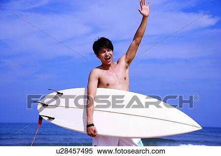 Stock Photography - Man holding the surfboard, waving one hand and smiling happily. Fotosearch