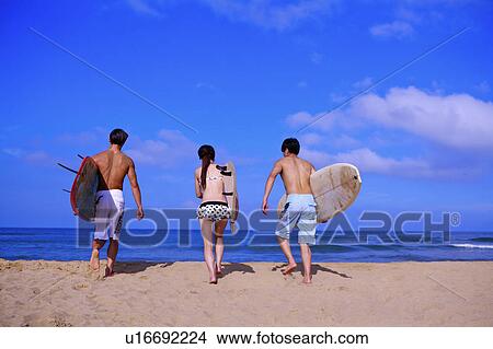 Picture - Three people holding the surfboards and walking toward the sea. Fotosearch