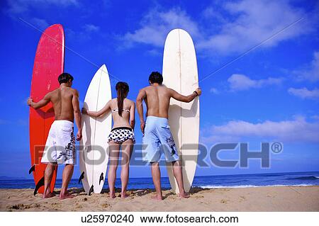 Stock Image - Three people holding the surfboards and standing on the beach, rear view. Fotosearch