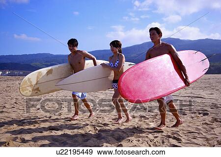 Stock Photo - Three people holding the surfboards and walking on beach. Fotosearch