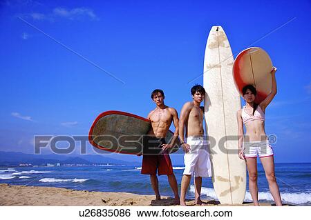 Stock Photograph - Three people standing on beach with surfboards. Fotosearch