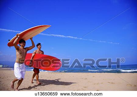 Stock Photo - Two men carrying surfboards and walking on beach. Fotosearch