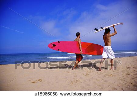Stock Photo - Two men carrying surfboards and walking on beach, rear view. Fotosearch
