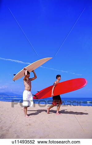 Stock Image - Two men carrying the surfboards and walking on beach. Fotosearch