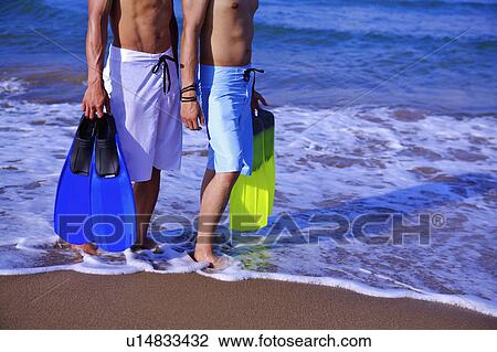 Stock Image - Two men holding the flippers and standing on beach. Fotosearch