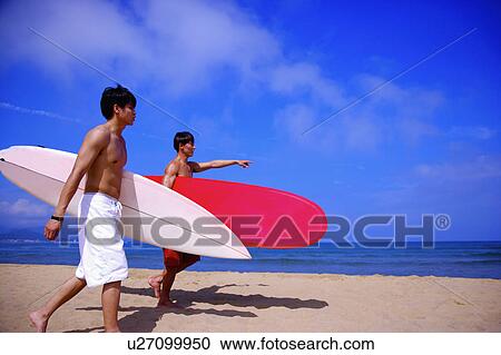 Stock Image - Two men holding the surfboards and walking on the beach. Fotosearch