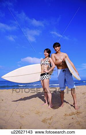 Stock Image - Two people holding the surfboards and standing on the beach. Fotosearch