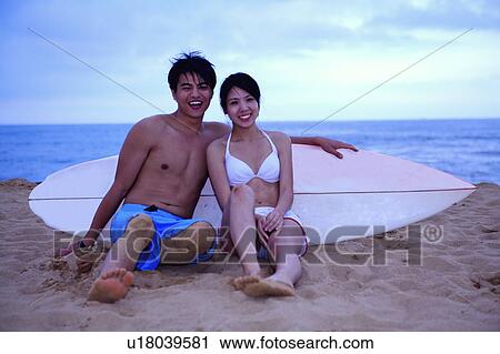 Stock Image - Young man and young woman sitting on beach with surfboard and smiling at the camera. Fotosearch