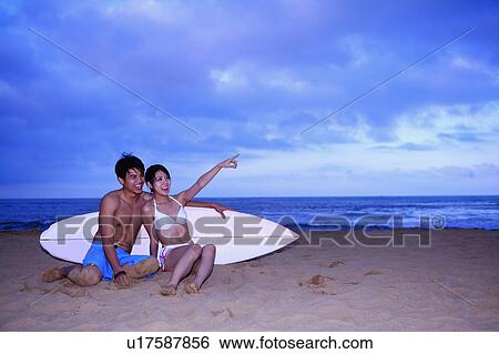 Young man and young woman sitting on beach with surfboard and looking away View Large Photo Image Stock Photograph - Young man and young woman sitting on beach with surfboard and looking away. Fotosearch