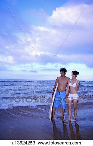 Stock Image - Young man and young woman standing on beach with surfboard and holding hands together. Fotosearch