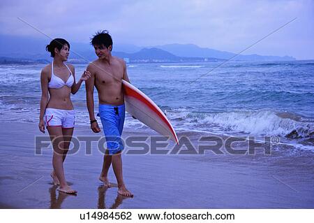 Stock Image - Young man and young woman walking on beach and holdinh a surfboard. Fotosearch