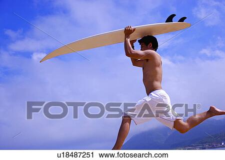 Stock Image - Young man carrying surfboard on head and jumping, side view. Fotosearch