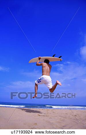 Young man carrying the surfboard on head and jumping in the mid-air, rear view View Large Photo Image Picture - Young man carrying the surfboard on head and jumping in the mid-air, rear view. Fotosearch