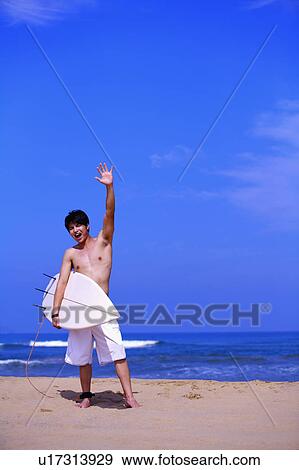 Stock Photo - Young man holding surfboard on beach and waving on hand. Fotosearch
