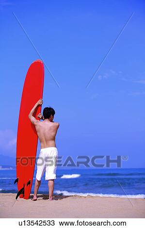 Young man holding the surfboard and standing on beach, rear view View Large Photo Image Stock Image - Young man holding the surfboard and standing on beach, rear view. Fotosearch