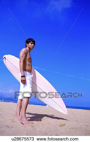 Stock Image - Young man holding the surfboard on beach and looking away. Fotosearch