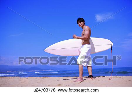 Stock Photo - Young man holding the surfboard on beach and looking at the camera. Fotosearch