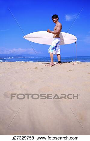 Stock Photo - Young man holding the surfboard, looking away and smiling. Fotosearch