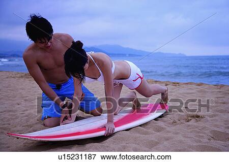 Stock Photo - Young man teaching young woman practicing on surfboard on beach. Fotosearch