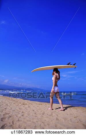 Stock Image - Young woman carrying a surfboard and walking on beach. Fotosearch