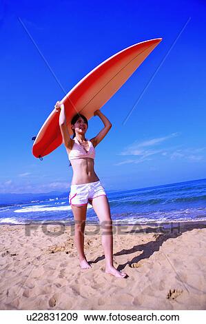 Stock Photo - Young woman carrying surfboard on head and walking on beach. Fotosearch