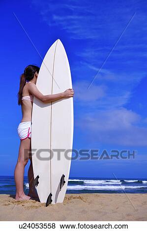 Stock Photo - Young woman holding surfboard on beach and looking away. Fotosearch
