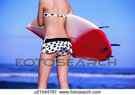 Stock Photo - Young woman holding the surfboad, rear view. Fotosearch
