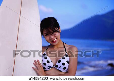 Stock Image - Young woman holding the surfboard and smiling at the camera. Fotosearch