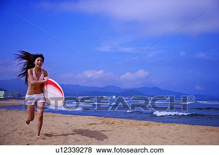 Stock Photo - Young woman holding the surfboard and running on beach. Fotosearch
