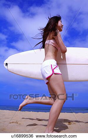 Stock Photograph - Young woman holding the surfboard and running on beach, side view. Fotosearch