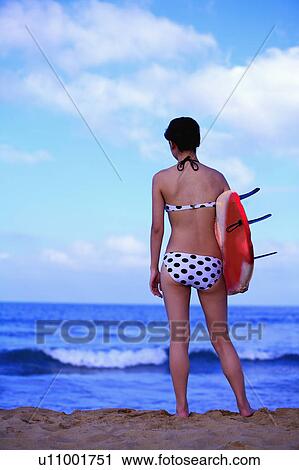 Stock Image - Young woman holding the surfboard on the beach, rear view. Fotosearch