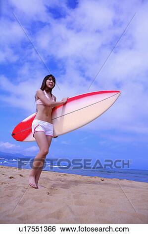 Stock Photograph - Young woman holding the surfboard on beach and looking away. Fotosearch