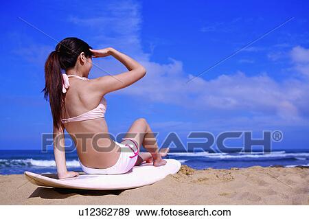 Stock Photo - Young woman sitting on surfboard and looking away, rear view. Fotosearch