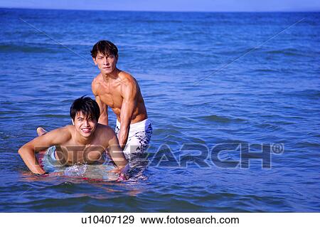 Stock Photo - Man helping young man surfing and looking at the camera together. Fotosearch