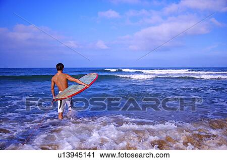Stock Image - Man standing in the water and holding the surfboard, rear view. Fotosearch