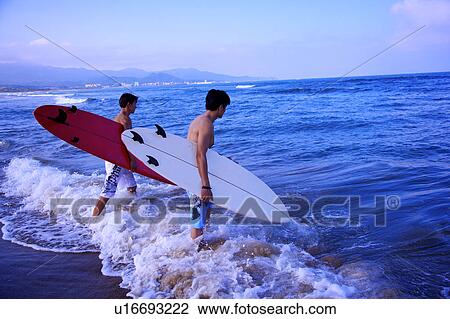 Stock Image - Two men holding the surfboards and walking toward the sea. Fotosearch