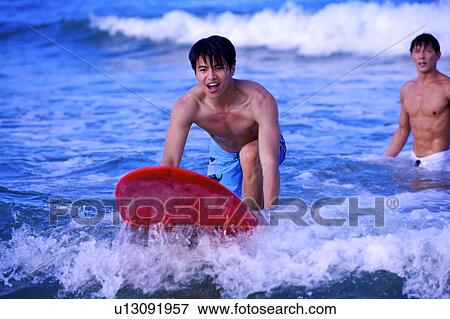 Stock Photo - Young man surfing on the sea and another man standing in the water. Fotosearch