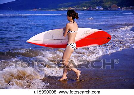 Stock Photo - Young woman holding the surfboard and walking toward the sea. Fotosearch