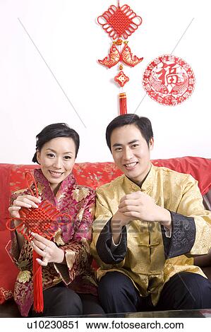 Stock Image - Young man with hands clasped and young woman holding a Chinese decoration, smiling at the camera together. Fotosearch