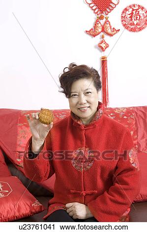 Stock Image - Mature adult woman in traditional clothing sitting on the sofa, holding a moon cake and smiling at the camera. Fotosearch