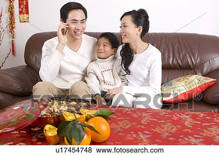 Stock Photo - Young couple with a child sitting on the sofa and smiling happily, on the phone. Fotosearch