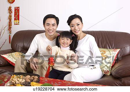Picture - Young couple with one child holding a tassel, sitting on the sofa and smiling at the camera. Fotosearch