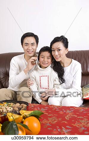 Picture - Young family with one child sitting on the sofa and smiling at the camera, on the phone. Fotosearch
