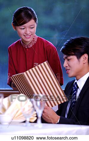 Stock Image - Young man looking at menu and telling young waitress what he wants. Fotosearch