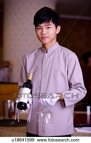 Stock Photo - Young waiter holding a bottle of red wine and two wine glasses, looking at the camera. Fotosearch