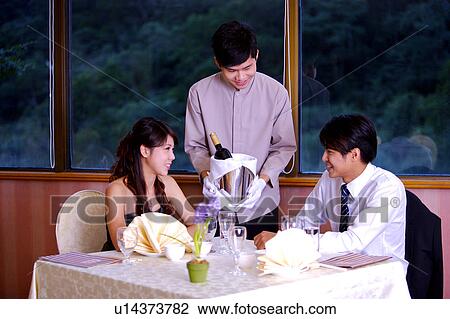 Stock Image - Young waiter holding a bottle of wine to two customer sitting beside the table. Fotosearch