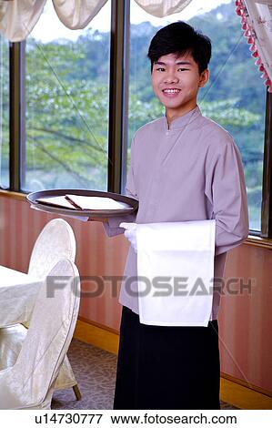 Stock Photo - Young waiter holding a tray and towel, smiling at the camera. Fotosearch