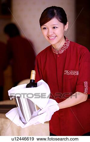 Stock Photo - Young waitress holding a bottle of red wine, looking away and smiling. Fotosearch