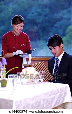 Picture - Young waitress writing the menu for young man who sitting near the table and looking at the menu. Fotosearch